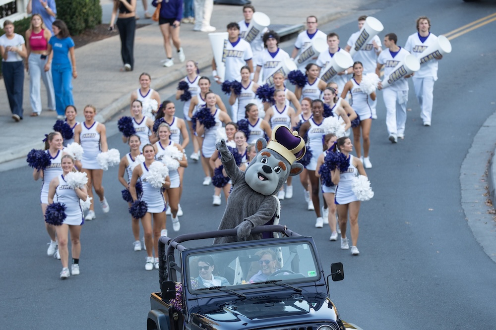 Duke Dog rides in an open-top vehicle, waving while a cheerleading squad and marching band members follow down a street during a parade.