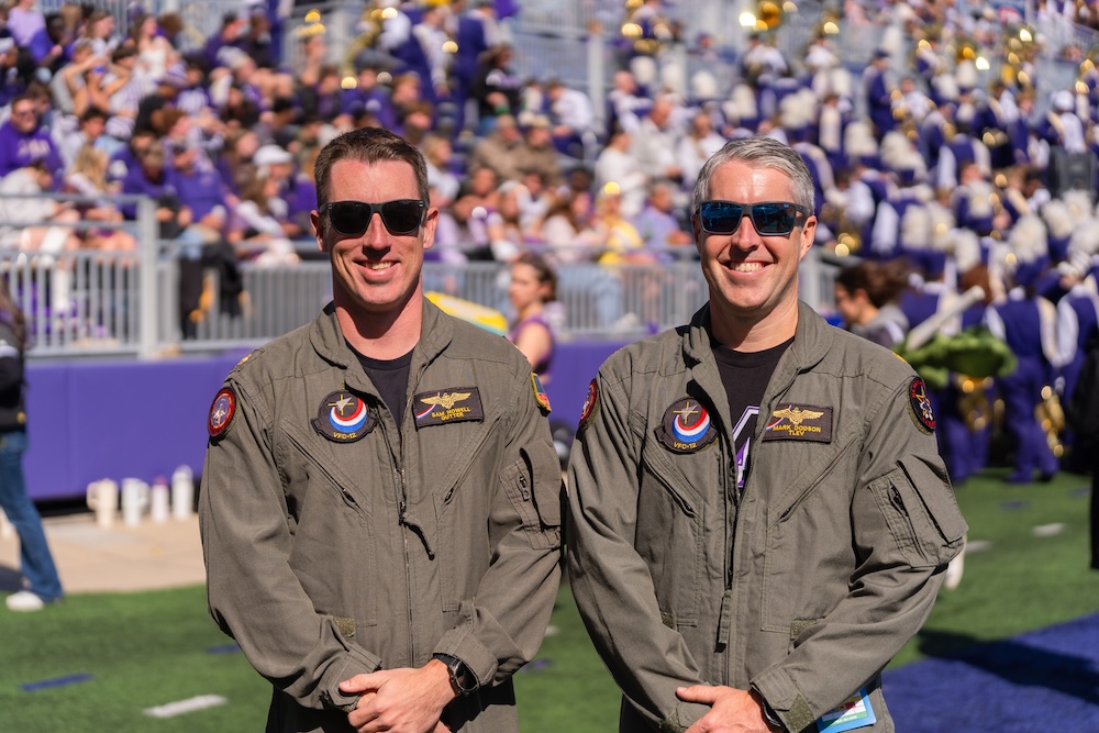 Two uniformed flight crew members wearing sunglasses stand on a football field, with a stadium crowd and marching band blurred in the background.