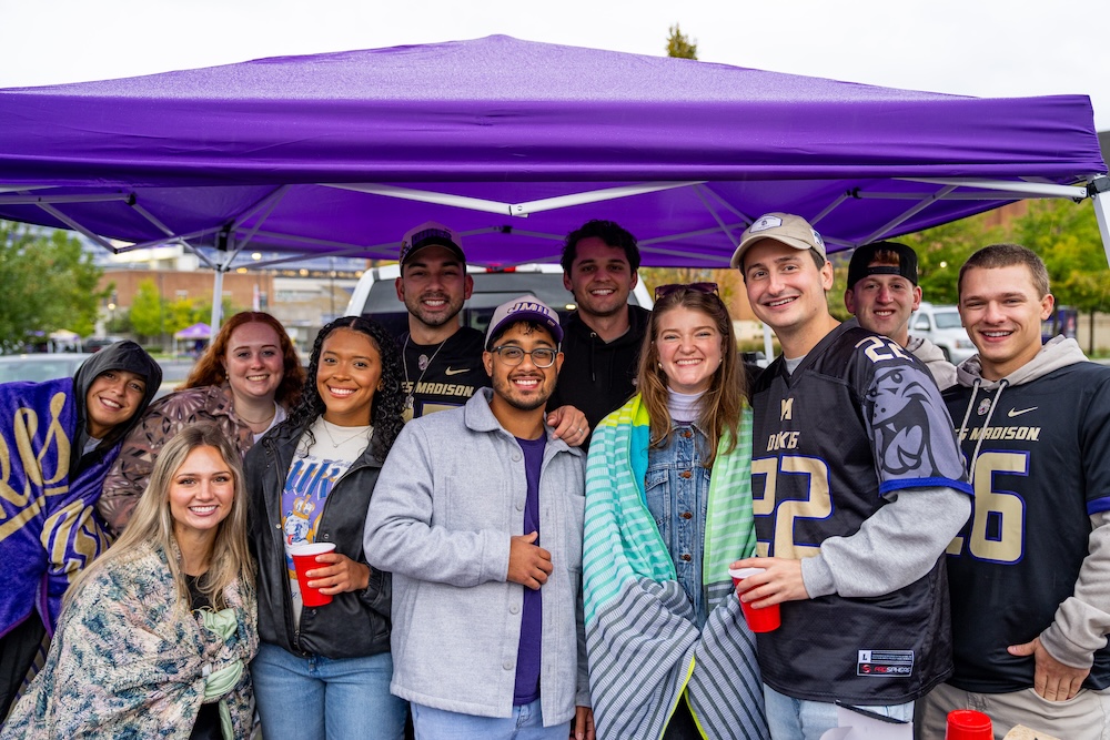 Group of people posing under a purple pop-up canopy at a tailgate, some wearing James Madison Dukes gear.