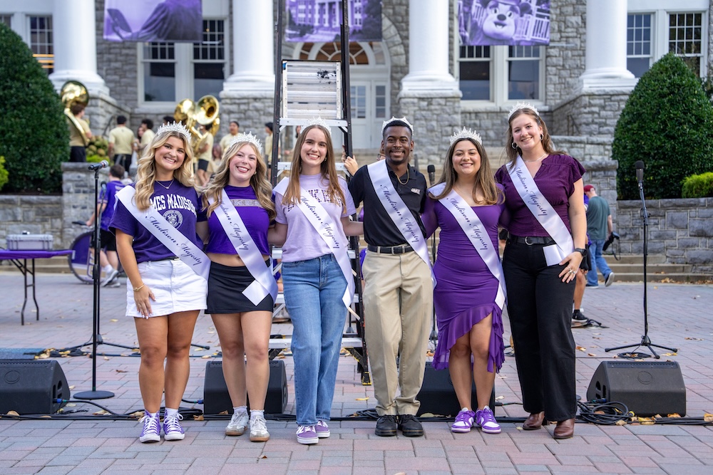 Group of six students wearing crowns and sashes pose in front of a campus building during an outdoor event.
