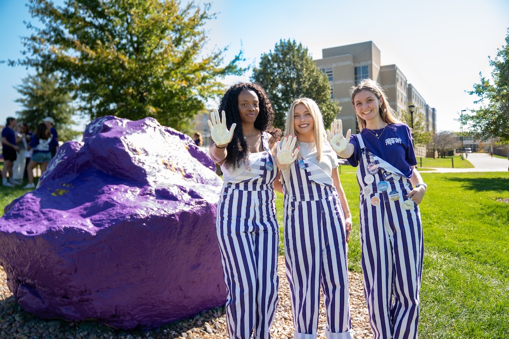 Three students in purple-and-white striped overalls stand beside a large purple painted rock on a sunny campus lawn, smiling and holding up paint-covered hands.