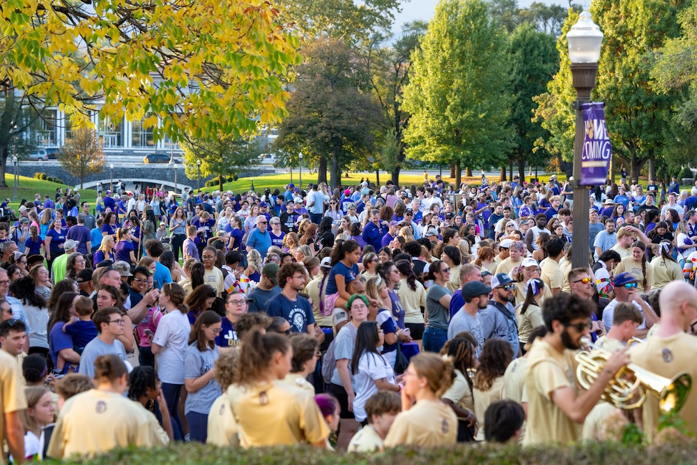 Large crowd gathered outdoors on a tree-lined campus walkway, with many people in school colors and a marching band in the foreground.