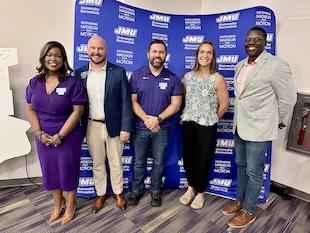 Five people pose smiling in front of a purple JMU-branded step-and-repeat banner at an indoor event.