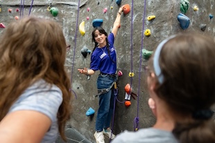 Climbing instructor wearing a harness gestures while standing on colorful holds on an indoor rock wall, with two students watching from the foreground.