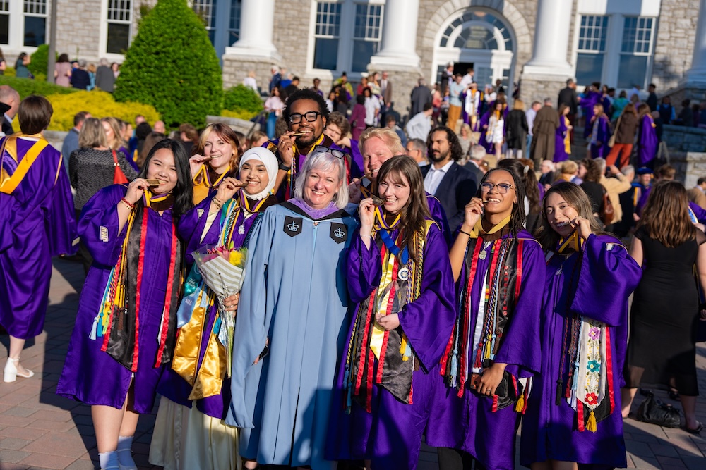 Group of graduates in purple gowns pose with a faculty member in front of a campus building, smiling and holding up their medals.