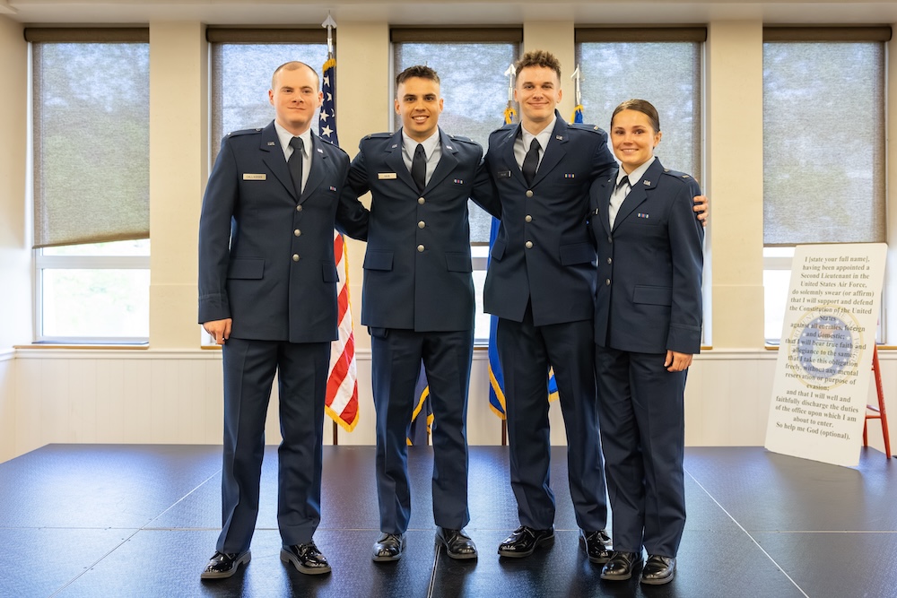 Four people during air force ROTC commissioning in matching military dress uniforms pose together on a stage indoors, with U.S. flags and large windows in the background.