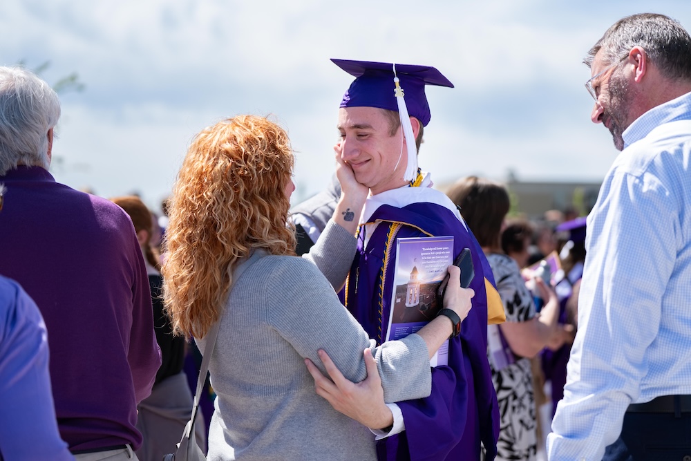 A graduate in a purple cap and gown smiles while being hugged by a woman in a crowd at an outdoor graduation ceremony.