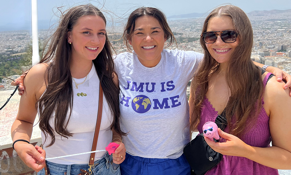 Dr. Finney, Mara and Riley travel around a street in Greece lined with white buildings with blue doors and pink flowers.