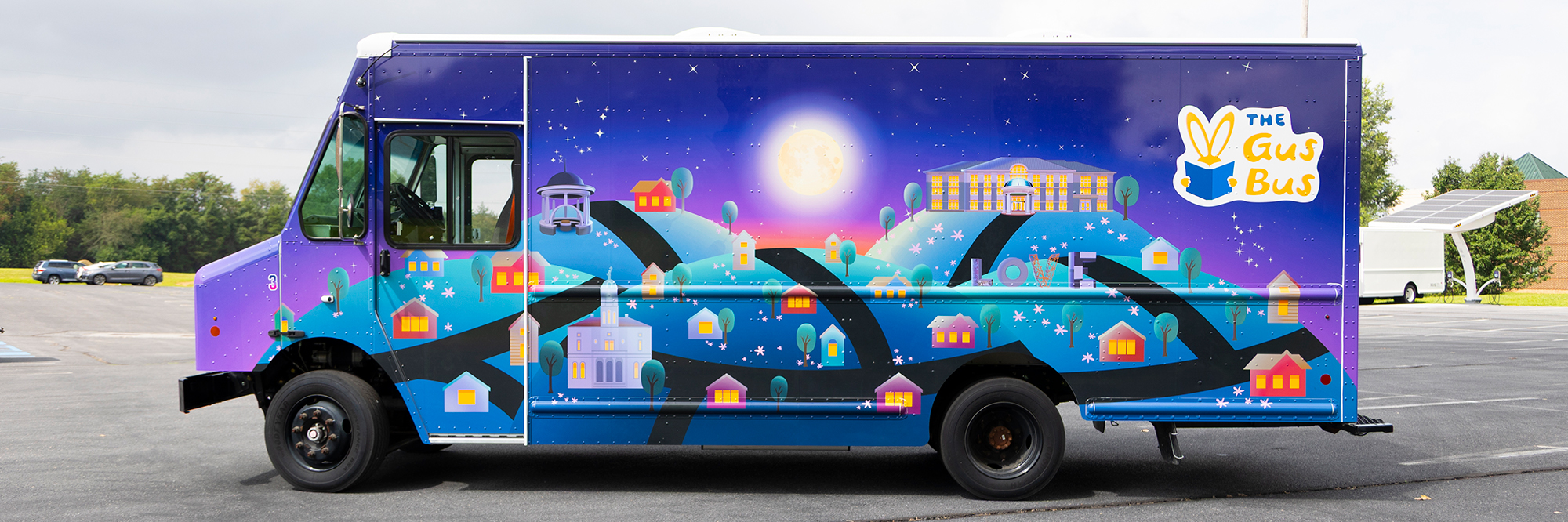 The side of the new Gus Bus vehicle, featuring a nighttime motif with deep purples and blues over Harrisonburg iconic locations, including the Court Square courthouse and the LOVE statue. 