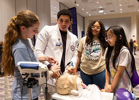A PA student demonstrates working with a patient on a simulation mannequin to three middle school students.