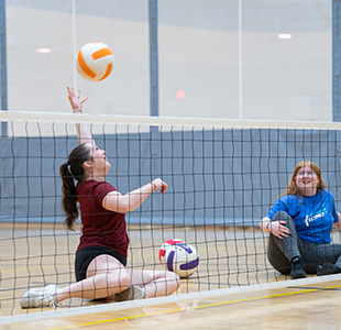 A student serves the ball in a round of sitting volleyball.