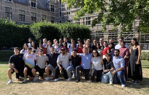 Large group of people posing together on a lawn in a courtyard, with historic stone buildings and leafy trees in the background.