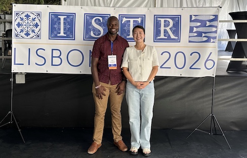 Two attendees wearing conference badges stand smiling in front of an ISTR banner that reads “LISBON 2026” inside a tented venue.
