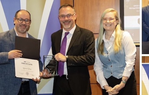 Three people smiling at an indoor awards presentation, with two holding a certificate folder and a glass trophy.