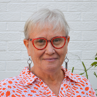 Michele. Woman with short white hair wearing red framed glasses.