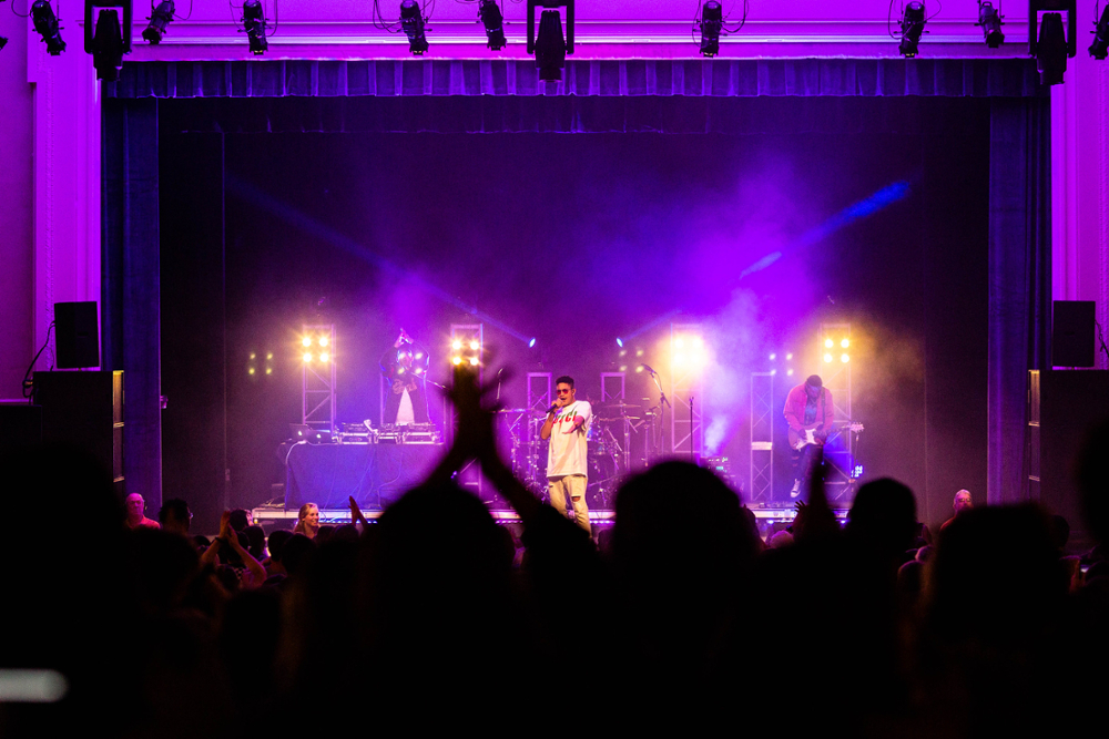 a view from the the audience as Bryce Vine performs at a concert at JMU, with the stage lit up with cool purple lights shine through haze from a fog machine