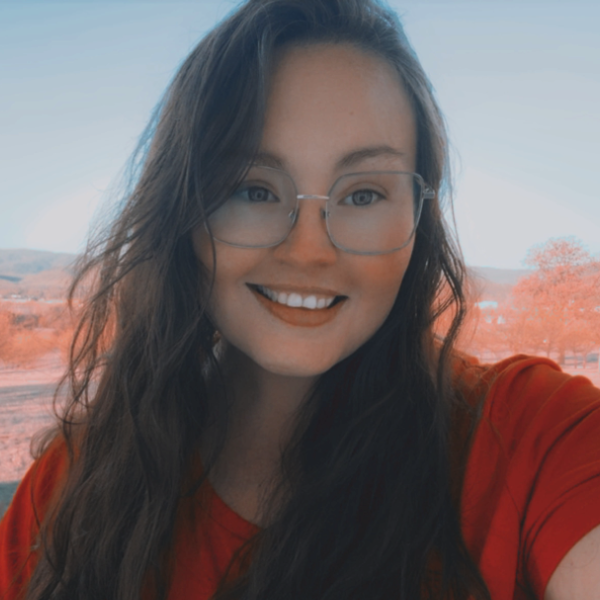 Danielle Wren with long dark hair and round glasses taking a selfie outdoors, with a softly blurred landscape in the background.