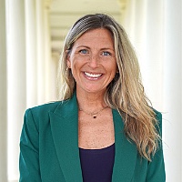 Smiling woman with long blonde hair wearing a green blazer, standing in a bright hallway.