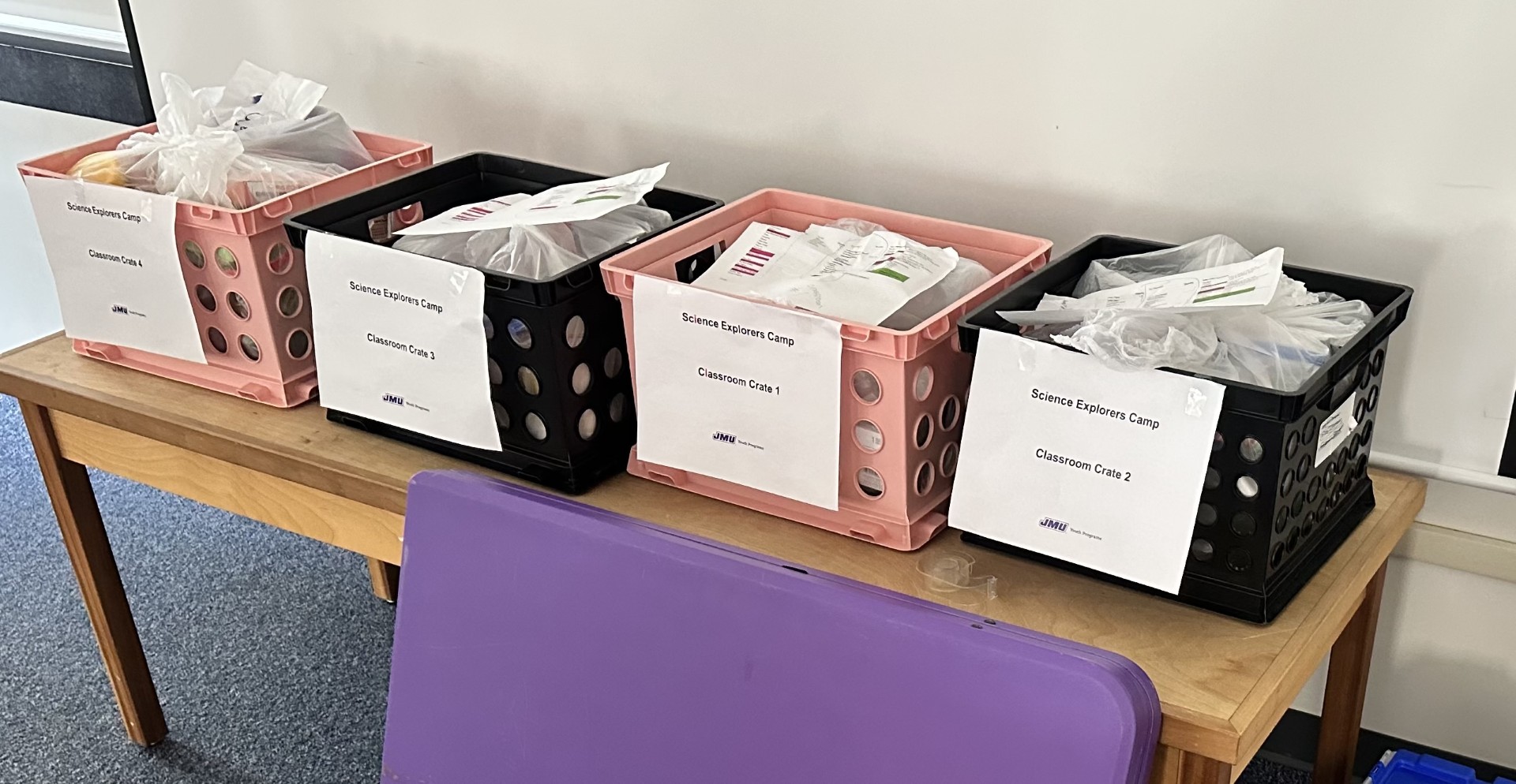 Four plastic storage crates labeled for “Science Explorers Camp” sit on a wooden table, filled with bagged supplies