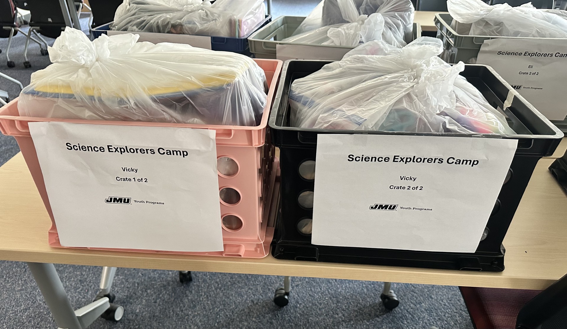 Plastic crates filled with bagged supplies on a table, labeled for a Science Explorers Camp (crate 1 of 2 and crate 2 of 2).