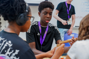 A focused student with a purple lanyard works with classmates on a hands-on project in a classroom setting.