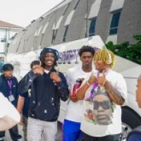 Three students smiling and posing near a tour bus, surrounded by a diverse group of people in an outdoor setting.