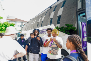 Three students smiling and posing near a tour bus, surrounded by a diverse group of people in an outdoor setting.