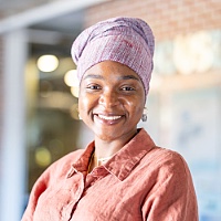 Musu, smiling and wearing a rust-colored shirt, standing outdoors with a softly blurred background.