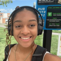 Smiling young woman wearing over-ear headphones and a backpack, taking a selfie outdoors near a sign.