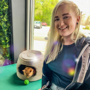 Youth Programs Director smiles while sitting beside a cat in a cozy pet house, with a green ball nearby.
