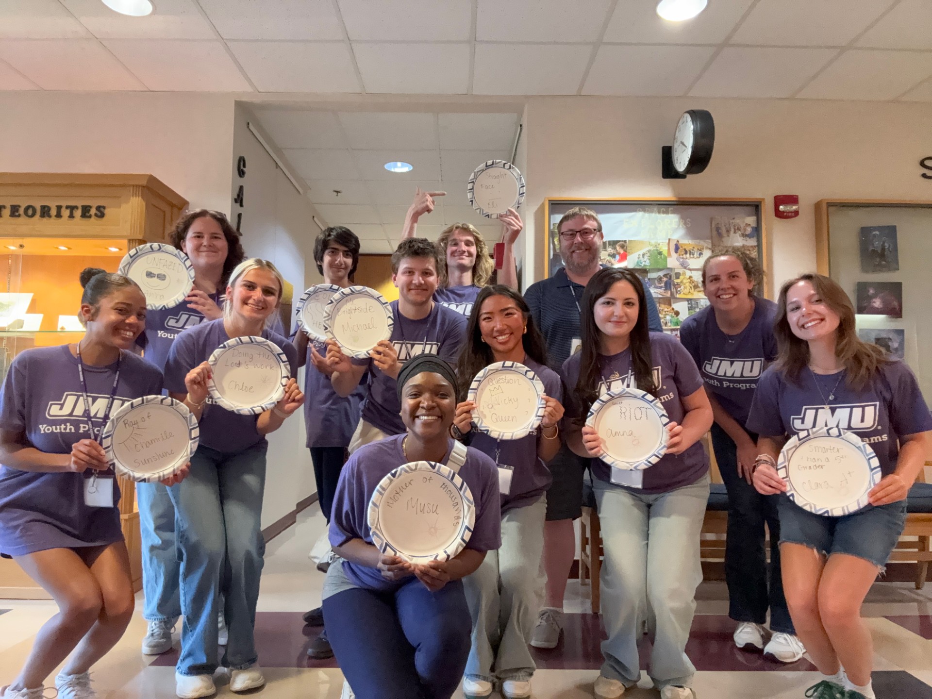 Group of counselors in matching purple JMU Youth Programs shirts pose indoors, smiling and holding paper plates with handwritten messages.