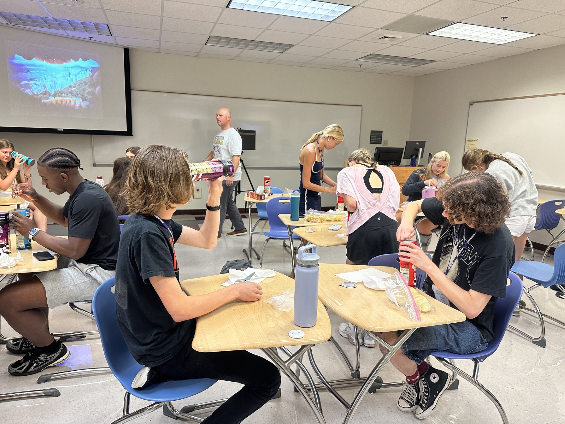 Students work at desks in a classroom, eating snacks and handling Pringles cans and other supplies while an instructor stands near a projector screen.