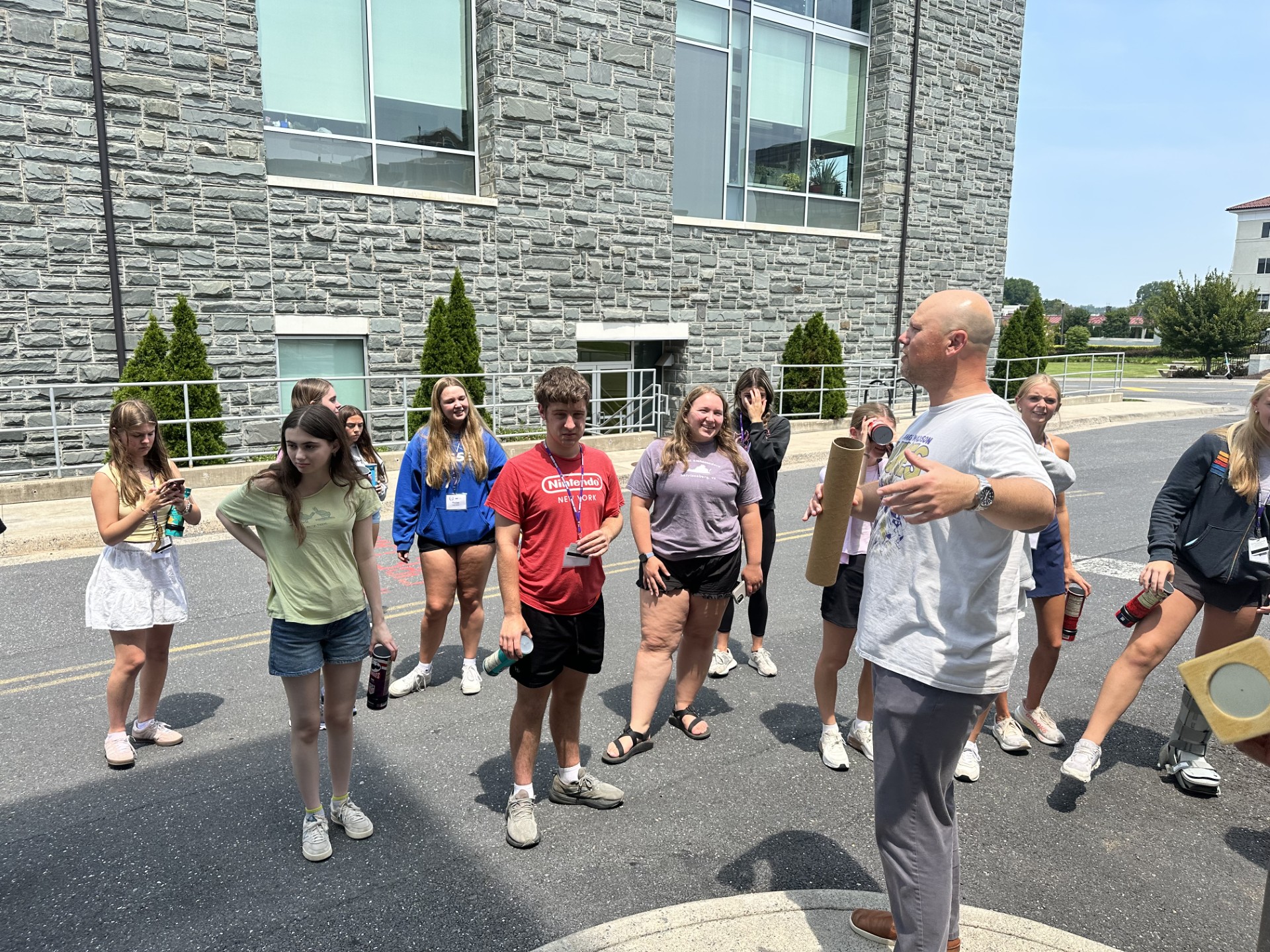 Group of teenagers stands outside a stone building while an adult speaks and gestures during an outdoor activity.