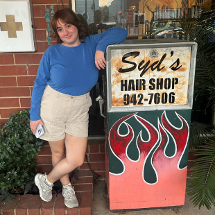 Youth Programs Coordinator in a blue sweatshirt and beige shorts leans against a vintage hair salon sign featuring flames, which reads "Syd's Hair Shop."