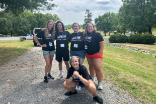 A group of five JMU student counselors wearing matching black t-shirts pose together outdoors, smiling for the camera.