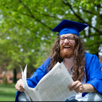 Zack, a joyful graduate in a blue cap and gown reads a newspaper while sitting outdoors among green trees.