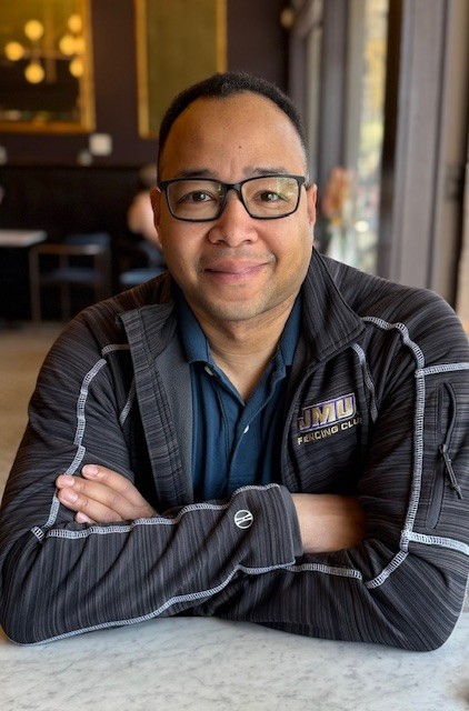 Fencing instructor wearing glasses and a JMU jacket smiles at the camera