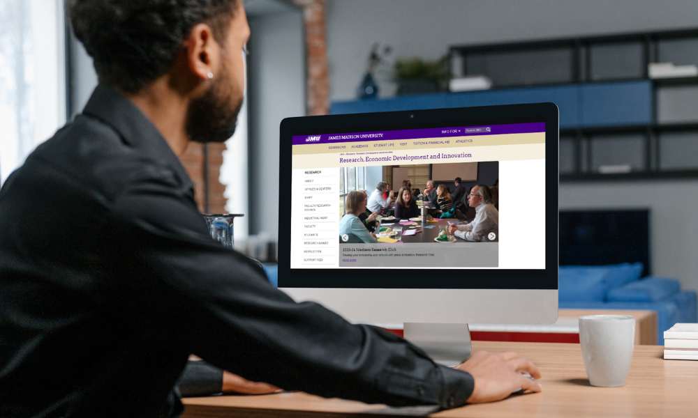 A man, sitting at a desk and looking at a computer monitor. The website for JMU's division of Research, Economic Development and Innovation is displayed on the computer's screen.