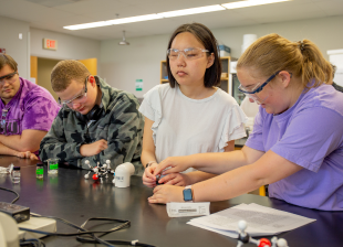 A group of students wearing safety goggles manipulating lab equipment while seated around a lab table