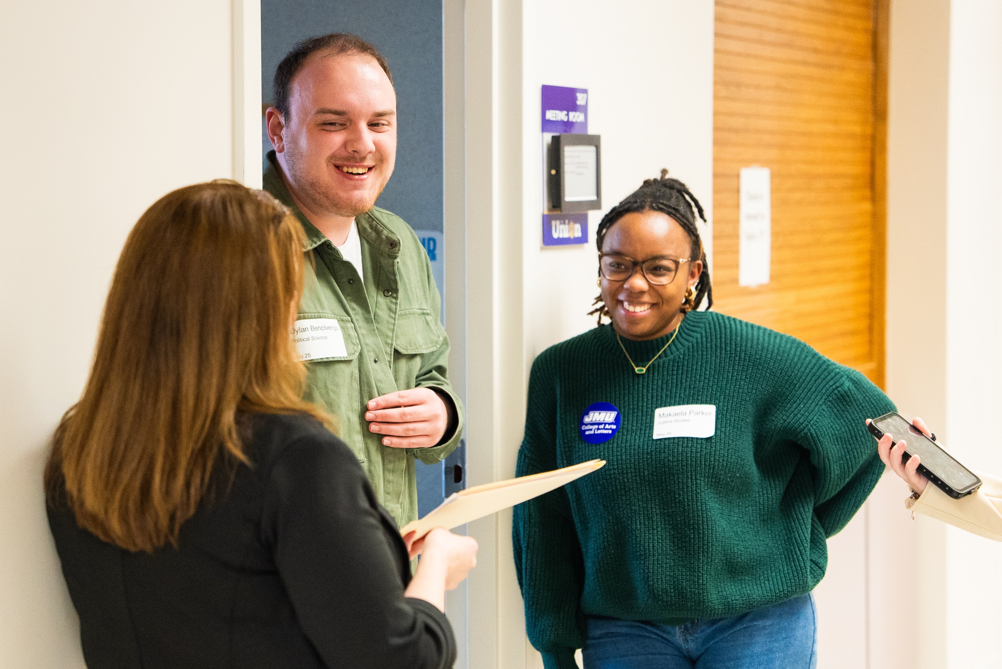 People engaging in conversation in a hallway, with smiling faces, and there is a person holding a folder.
