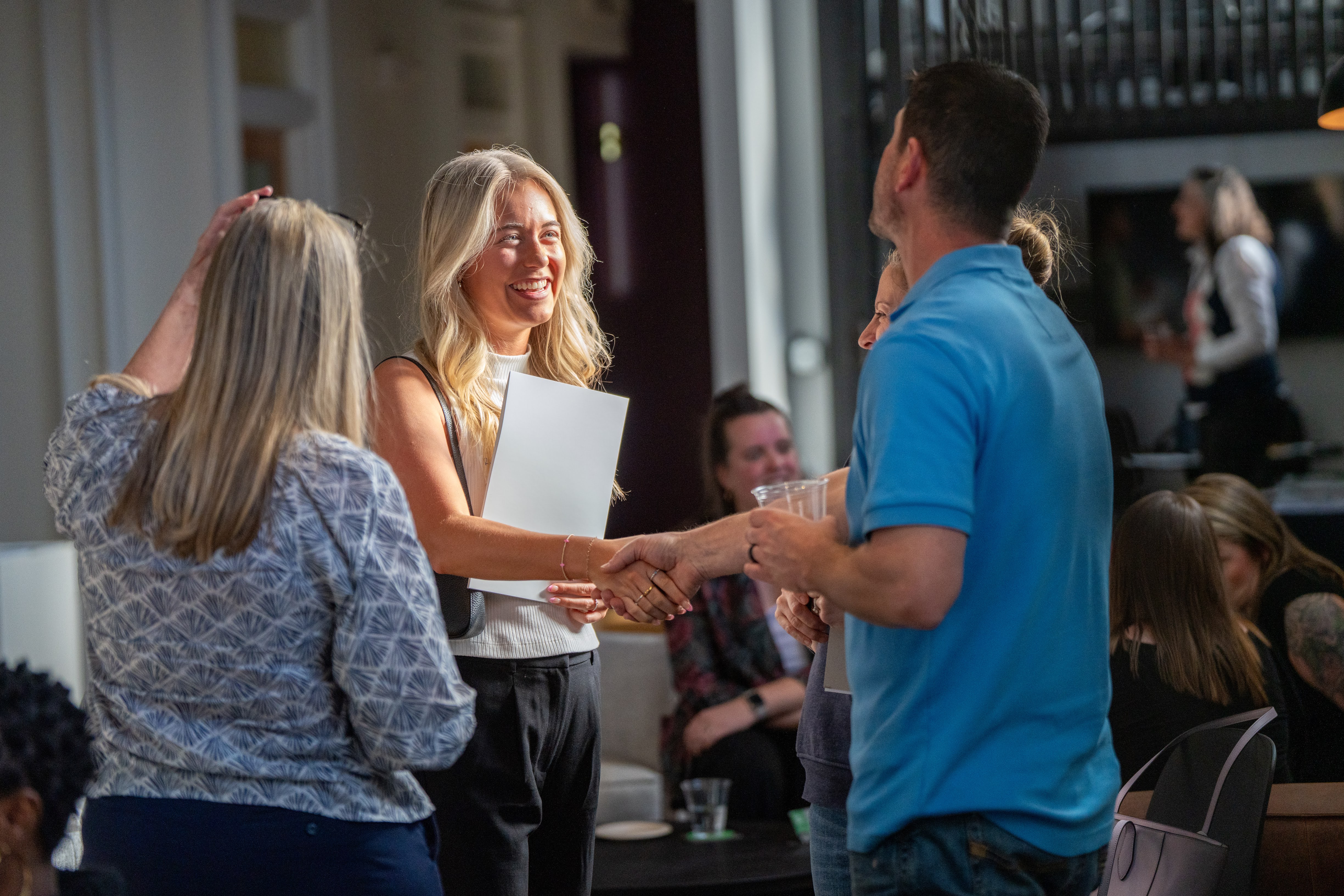 A woman with blonde hair smiles while shaking hands with a man in a blue shirt, surrounded by a group of people engaged in conversation at a social event.