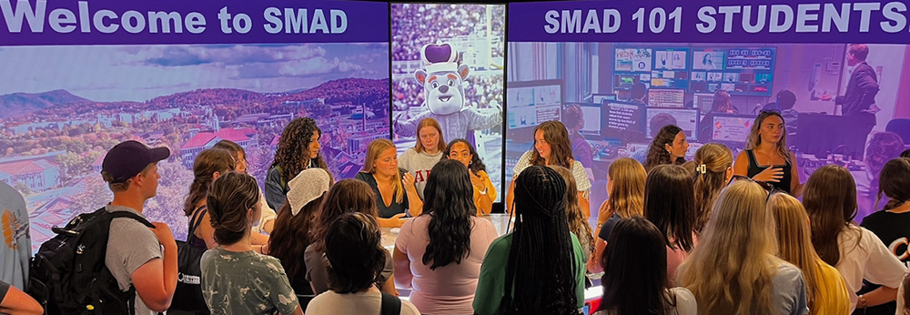 A group of people listen to speakers at a news desk in front of a studio LED wall that says "Welcome to SMAD" and shows the Duke Dog.