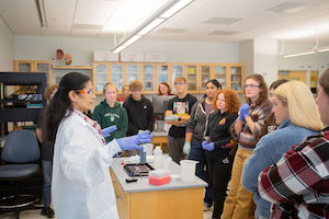 A lab instructor in a white coat and gloves demonstrates equipment at a workbench while a group of students gathers around watching in a classroom laboratory.