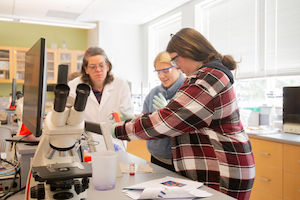 Three researchers in a laboratory review samples and notes next to a microscope and lab equipment.