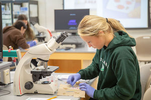 Student in a green hoodie and gloves works at a lab bench beside a microscope in a classroom science lab.