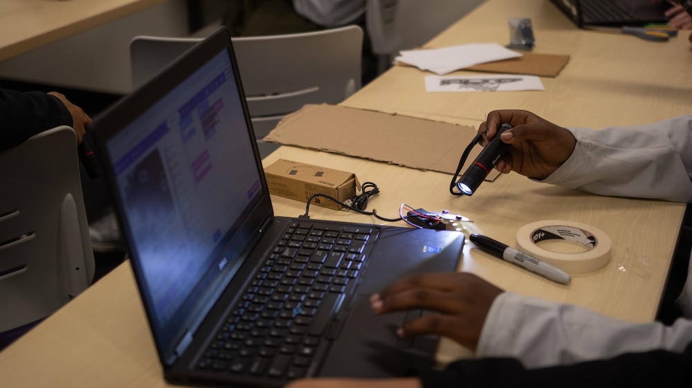 A person tests a small electronic circuit with a flashlight beside an open laptop on a classroom table.