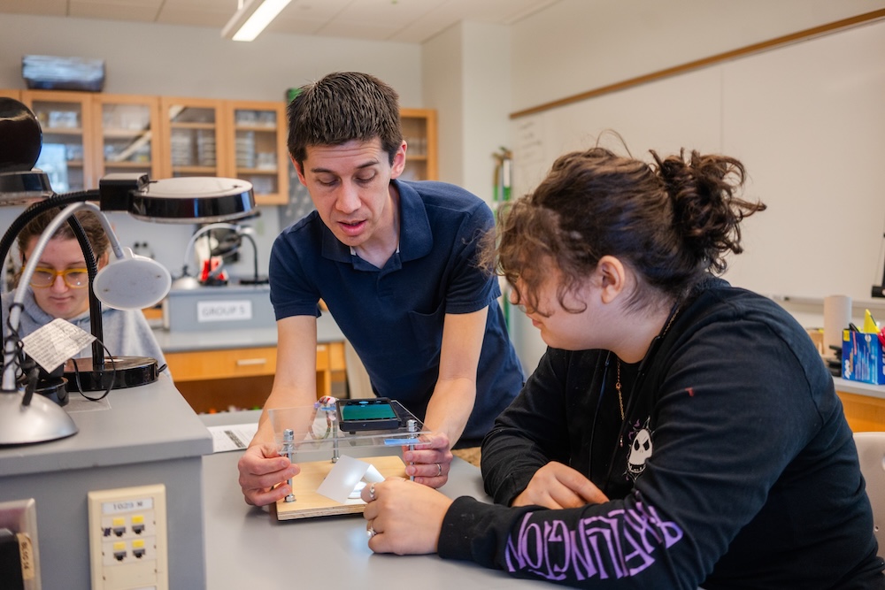 Teacher demonstrates a small science setup on a lab table while a student watches in a classroom laboratory.