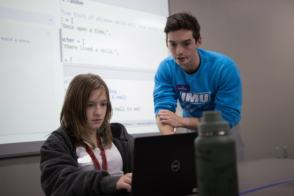 Instructor leans over to help a student working on a laptop in a classroom, with a projected coding screen in the background.