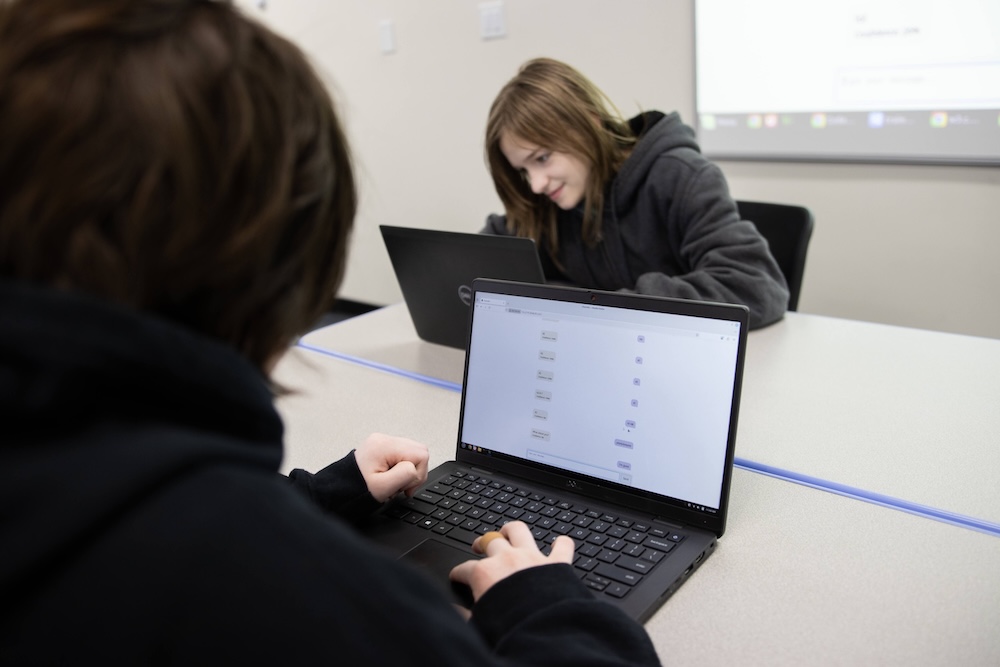Two students working on laptops at a classroom table, with a projected screen in the background.