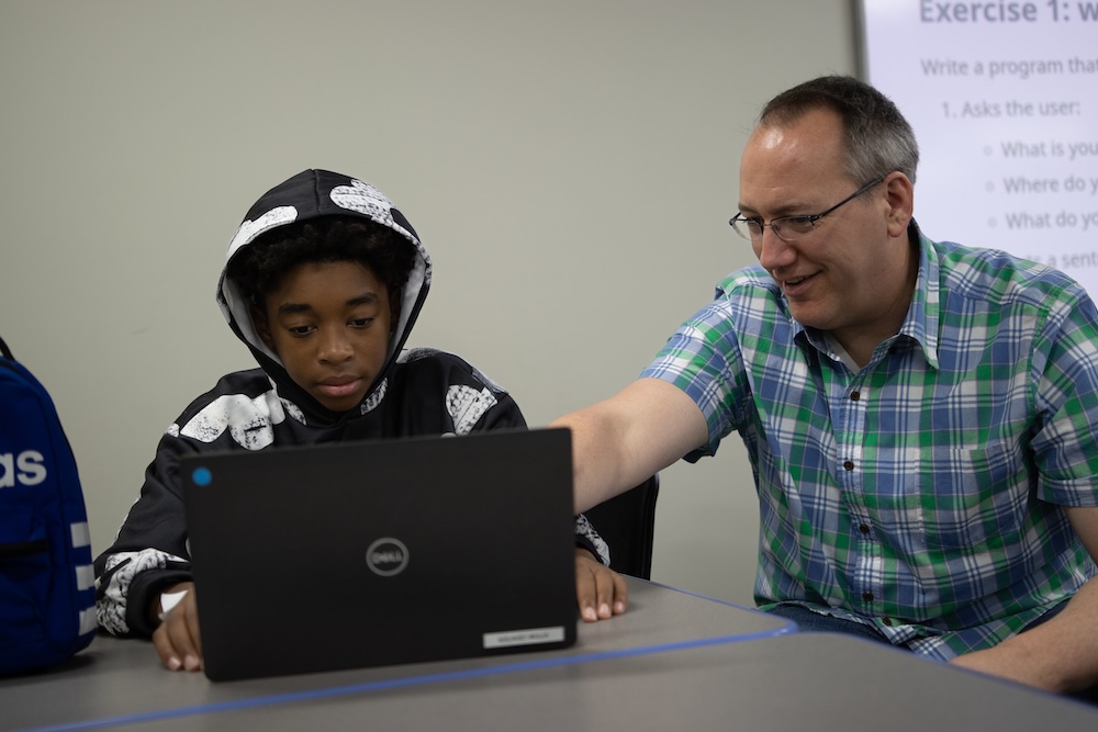 An adult helps a student use a laptop at a classroom table, pointing at the screen while they work together.